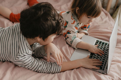 High angle view of brother and sister lying  on bed at home with laptop 