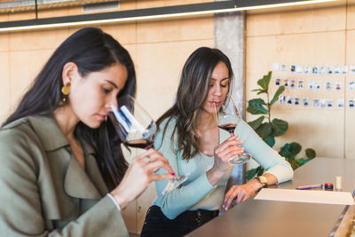 Female sommeliers in smart casual clothes enjoying aroma of red wine while sitting at counter during degustation session in bar with bartender