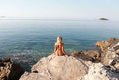 Rear view of naked woman sitting on cliff by sea