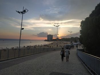 People on street by sea against sky during sunset