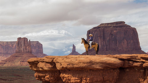 Full length of man sitting on horse over rocky cliff against sky