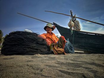 Senior fisherman sitting by ropes and boat at sandy beach