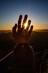 Cropped hand of man against sun during sunset