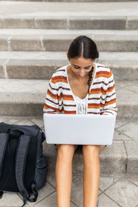 High angle of young female student browsing netbook and doing homework while sitting on concrete steps near backpack in city