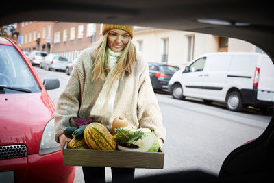 Smiling woman with freshly produce vegetables in container standing near car trunk