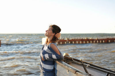 Brunette girl in striped sweater, bare shoulder and denim skirt, smiling near an old gray boat 