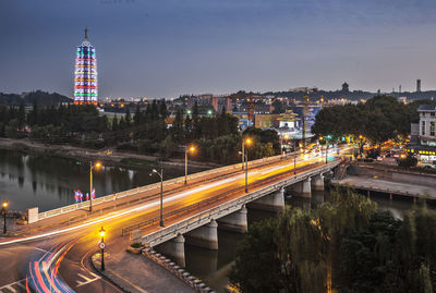 Light trails on road by illuminated buildings against sky at night