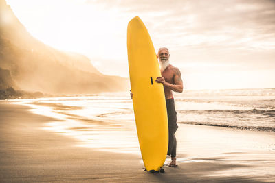 Portrait of man with surfboard standing on beach by sea against sky