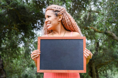 Young woman holding blackboard against trees