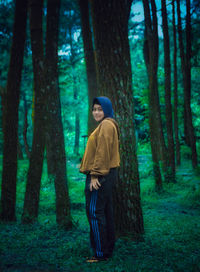 Full length side view of young woman standing by tree trunk in forest