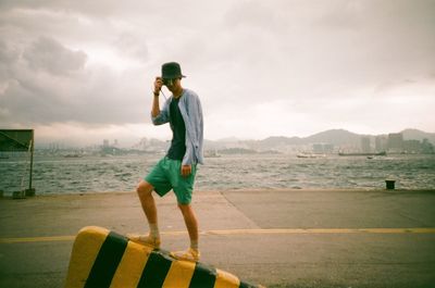 Full length of man with camera standing on retaining wall against river and cloudy sky