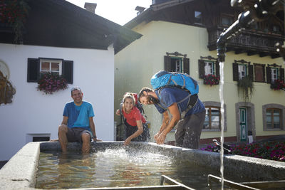 Happy hiker sitting by playful friends splashing water at fountain in village, mutters, tyrol, austria