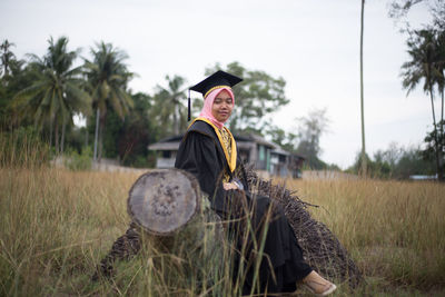 Young woman wearing graduation gown sitting on field
