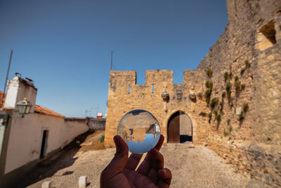 Person holding glass ball by building against sky