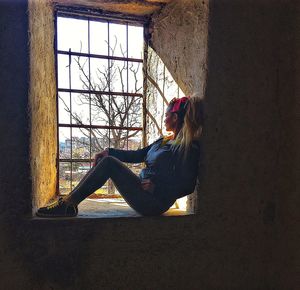 Side view of young woman sitting on window at home