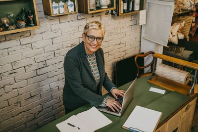 High angle portrait of smiling senior entrepreneur using laptop at home decor shop