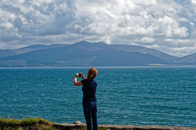 Rear view of woman photographing by lake against mountains and cloudy sky