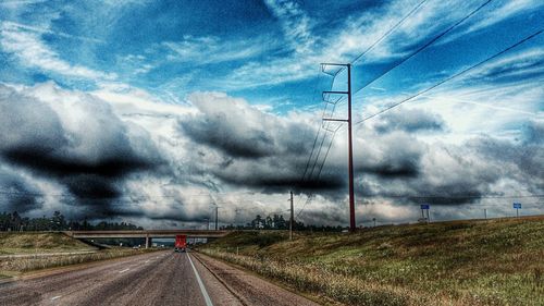 Electricity pylons on road against cloudy sky