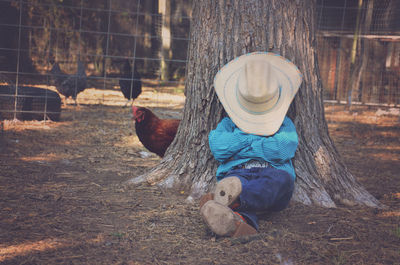 Cowboy relaxing with hat over face in farm against tree trunk