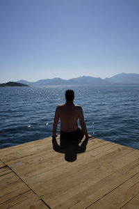 Rear view of man sitting on shore against clear sky