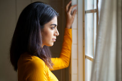 Side view of young woman looking through window at home