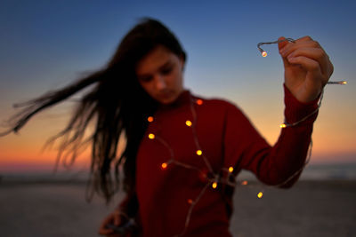 Woman holding illuminated string light while standing at beach during sunset