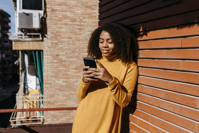 Young woman looking through smart phone while standing against brick wall