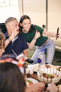 Teenage girl taking selfie with grandfather on mobile phone while having food at campsite