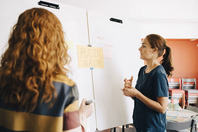 Confident female colleagues discussing plastic management project over whiteboard at creative office