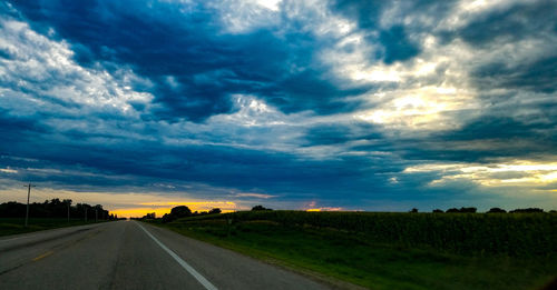 Road passing through field against storm clouds