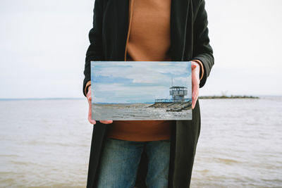 Midsection of woman holding painting against sea and sky