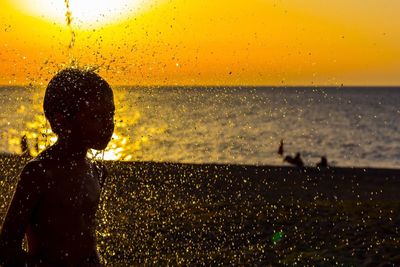 Water splashing on silhouette boy at beach against sky during sunset