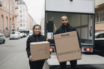 Portrait of male and female workers carrying boxes while standing against delivery van in city