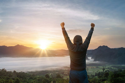 Rear view of woman with arms raised standing against sky during sunset