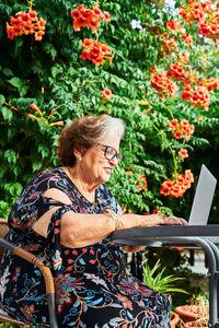 Concentrated elderly lady in casual outfit and eyeglasses sitting in chair at table while surfing on netbook near cup on saucer and green plants with flowers in yard in daylight