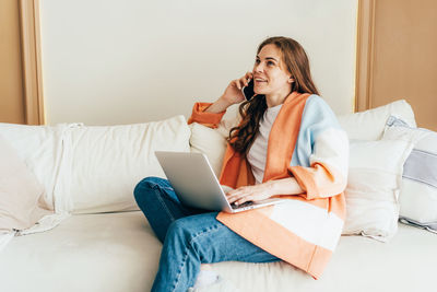 Young business woman smiling and talking on phone while work with laptop at home sitting on sofa.