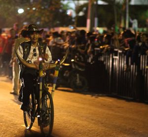 Man wearing cowboy hat riding bicycle on street at dusk during event