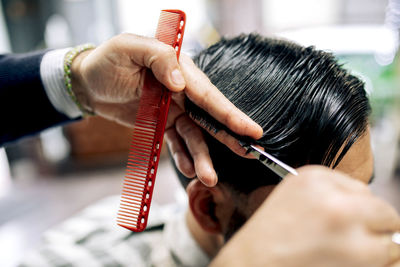 Crop anonymous barber using scissors and comb to cut hair of male client while working in grooming salon