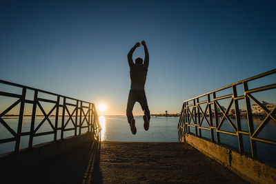 Full length of man jumping over pier at beach against sky during sunset