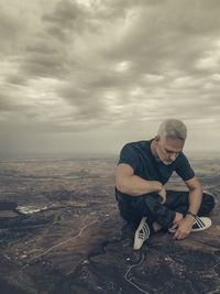 Full length of mature man sitting on rock against cloudy sky