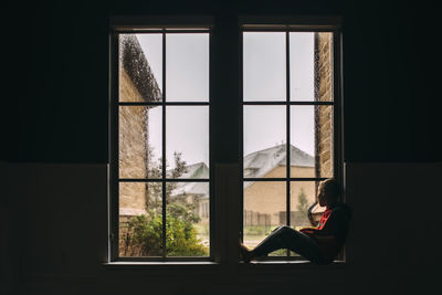 Side view of boy looking through window while sitting at home during rainfall