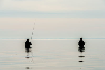 Rear view of men fishing while standing in sea against sky