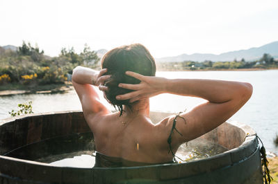 Back view of thoughtful unrecognizable female doing hair bun while chilling in barrel with hot water and seaweed and admiring calm lake in morning