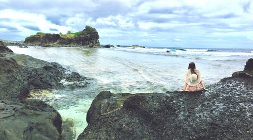 Rear view of woman sitting on rock by sea against cloudy sky