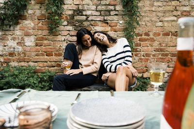 Happy female friends enjoying together while sitting on bench against wall at dinner party