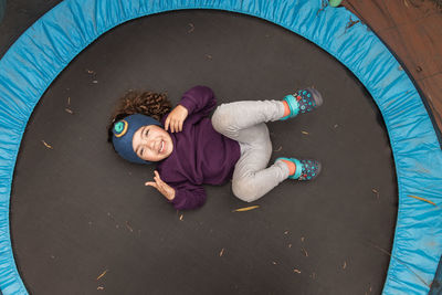 Top view full body of joyful ethnic child looking at camera while lying on trampoline and having fun at daytime