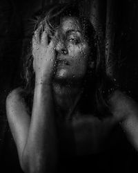 Close-up portrait of woman seen through wet window against black background