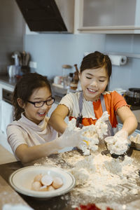 Smiling sisters with messy hands kneading dough in kitchen at home