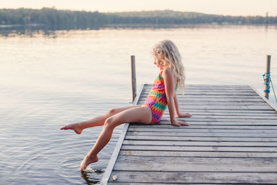 Caucasian blonde girl child sitting on wooden dock pier by lake. pensive kid in swimsuit splashing 