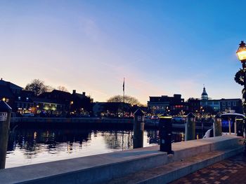 Illuminated buildings by river against sky at dusk